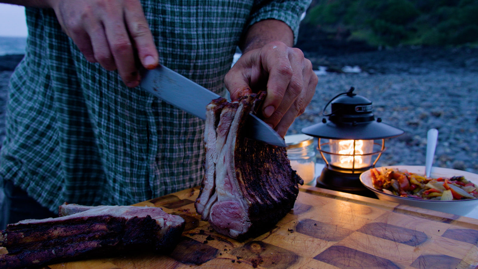 Slicing a fire-roasted bone-in rib of beef on a wooden board by the coast at dusk
